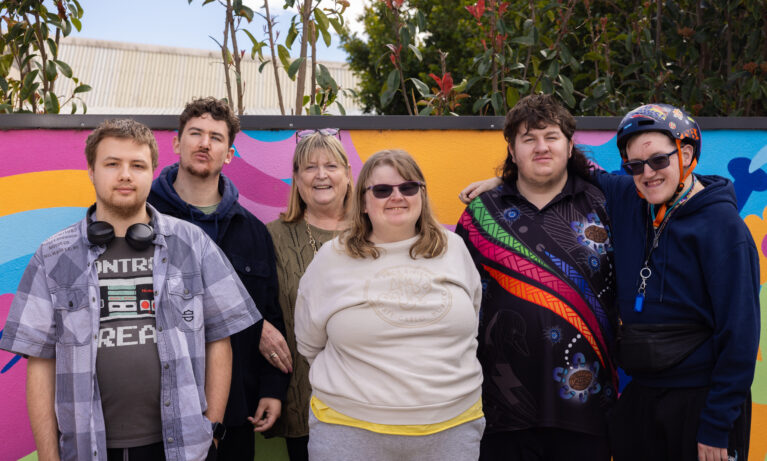 Group Support clients smiling and standing in front of a mural with a Support Worker.